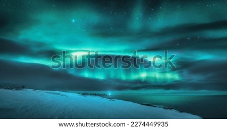 Aurora borealis over ocean. Northern lights and frozen sea coast. Starry sky with polar lights and clouds. Night winter landscape with aurora, sea with blurred water, snowy mountains. Travel