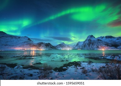 Aurora borealis over the sea coast, snowy mountains and city lights at night. Northern lights in Lofoten islands, Norway. Starry sky with polar lights. Winter landscape with aurora reflected in water - Shutterstock ID 1595659051