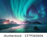 Aurora borealis over the sea, snowy mountains and city lights at night. Northern lights in Lofoten islands, Norway. Starry sky with polar lights. Winter landscape with aurora, reflection, sandy beach 