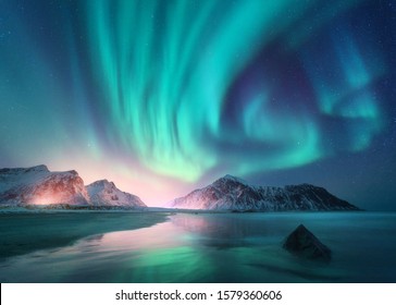 Aurora borealis über dem Meer, schneebedeckte Berge und Lichter der Stadt nachts. Nordlichter auf den Lofoten-Inseln, Norwegen. Sternenhimmel mit Polarlichtern. Winterlandschaft mit Orora, Reflexion, Sandstrand 