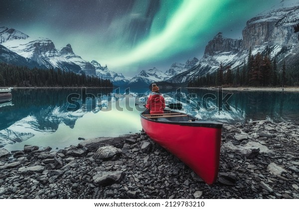 Aurora borealis over Spirit island with female\
traveler sitting on red canoe on Maligne Lake at Jasper national\
park, AB, Canada