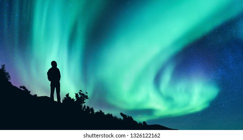 Aurora Borealis And Silhouette Of A Woman With Backpack At Night. Girl On The Hill, Starry Sky With Northern Lights. Sky With Stars And Polar Lights. Trekking. Landscape With Bright Aurora And People