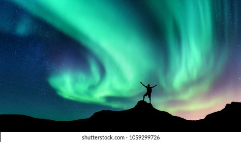 Aurora and silhouette of standing man with raised up arms on the mountain in Norway. Aurora borealis and happy man. Starry sky, green polar lights. Night landscape. Northern lights. Travel and tourism - Shutterstock ID 1059299726