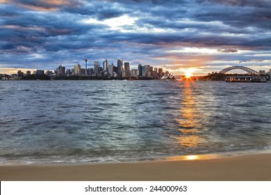 Australia Sydney City CBD And Harbour Bridge Landmarks At Sunset With Sun Touching Horizon And Blurred Harbour Waves On Sandy Beach 