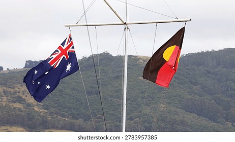 Australian and Aboriginal flags flutter from a white mast against a hilly landscape.