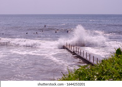 Australian Beach With Some Waves On It And Some People Practicing Water Sports. IN The Image We Can See A Wave Hitting Against The Rock Pool While Some Surfers Are Waiting For A Wave.