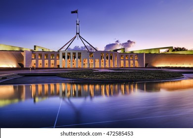 Australian national parliament house in Canberra. Facade of the building brightly illuminated and reflecting in blurred water of fountain pond under waving national flag on flagpole at sunset.
