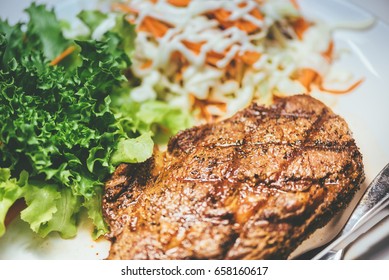 Australian Ribeye Beef Steak With Distinct Cross Char Marks. Knife And Fork On Side. On White Plate. Served Alongside Fresh Green Oak And Sliced Coleslaw.