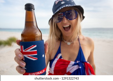 Australian Woman On The Beach Holding Or Offering A Beer And Cheerfully Smiling.  Focus To Bottle Which Is In A Foam Cooler Printed With The Australian Flag.   Aussie Culture, Party, Bbq