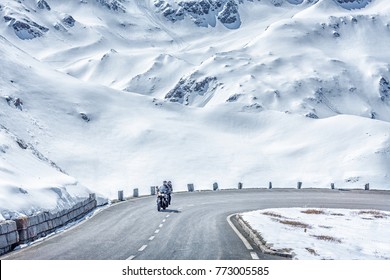 Austria, Tyrol, High Alpine road. Snowy scenery. Couple traveling by motorcycle, moving on speed by road curve of Grossglockner Hochalpenstrasse at snowy Alps mountains background. Sunny May day.