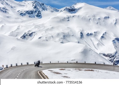 Austria, Tyrol, High Alpine road. Snowy scenery. Family traveling by motorcycle, moving on speed by road curve of Grossglockner Hochalpenstrasse at snowy Alps mountains background. Sunny May day.