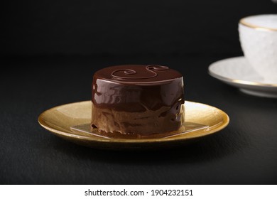 Austrian Cake, Sackertorte. Chocolate Cake On A Golden Plate. White Cup With Cappuccino. Side View Of A Chocolate Dessert. Black Textured Table.