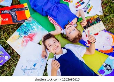 Authentic artist children in blue aprons girl and boy paints tickle each other by paint brushes, lying in creative chaos palette watercolor paints, brush, drawings.Painting outdoor. Top view