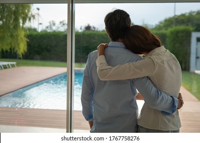 Authentic Back View Shot Of Lovely Embraced Married Couple Is Enjoying Time Together While Looking Through A Window With A View On A Garden With Pool At Home.