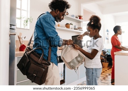 Authentic black family starts their day together in the morning, as the father helps his young daughter pack her school bag. It's a heartwarming depiction of fatherhood and family routine.