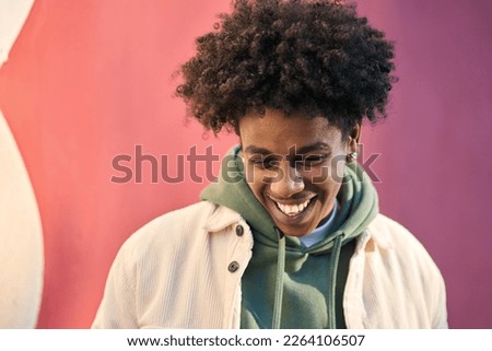 Authentic candid shot of young happy African American cool teen guy laughing on red wall lit with sunlight. Smiling cool rebel gen z teenager model standing outdoors having fun, portrait outdoors.