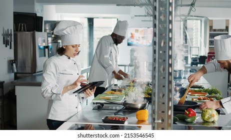 Authentic Chef Looking At Food Recipe On Digital Tablet Screen, Using Online Internet Website To Make Gastronomy Dish. Woman Cook With Device Making Professional Cuisine Restaurant Meal.