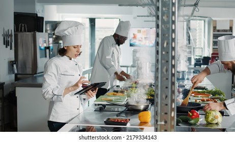 Authentic chef looking at food recipe on digital tablet screen, using online internet website to make gastronomy dish. Woman cook with device making professional cuisine restaurant meal. - Shutterstock ID 2179334143