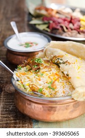 Authentic chicken biryani served with naan bread, fragrant pilau rice and yoghurt, in a metal pot.