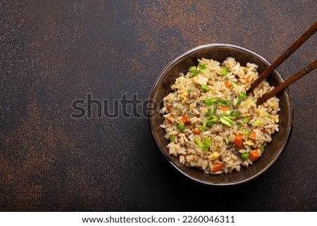 Authentic Chinese and Asian fried rice with egg and vegetables in ceramic brown bowl top view on dark rustic concrete table background. Traditional dish of China with space for text