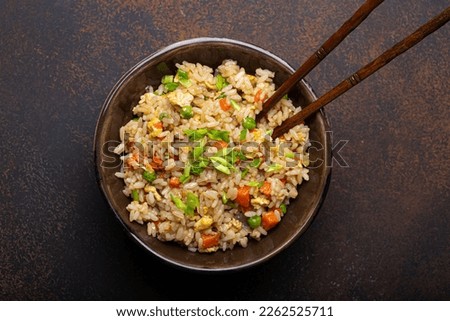 Authentic Chinese and Asian fried rice with egg and vegetables in ceramic brown bowl top view, dark rustic concrete table background. Traditional dish of China