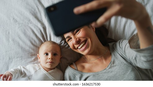 Authentische Nahaufnahme der Neumutter und ihres Neugeborenen, die einen Selbstgespräch oder einen Videoanruf zu Vater oder Verwandten in einem Bett führen. Konzept der Technologie, der neuen Generation, der Familie, der Verbindung, der Elternschaft, der Authentizität
