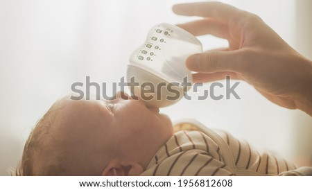 Authentic Close Up Shot of a Newborn Drinking Milk from Small Baby Bottle while Lying on the Back in Child Crib. Portrait of Caucasian Neonate Toddler at Home. Concept of Childhood and Parenthood.