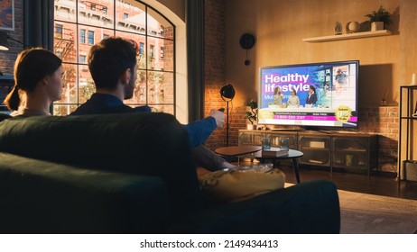 Authentic Couple Spending Time at Home, Sitting on a Couch and Watching TV with Big Flat Screen Display in Their Stylish Loft Apartment. Man and Woman Streaming Reality Show or Infomercial.