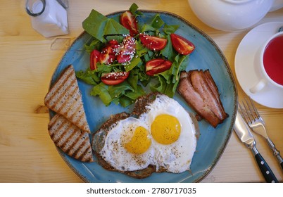 Authentic flat lay composition of a hearty morning meal. A rustic blue plate features crispy sunny-side-up fried eggs, bacon strips, grilled toast, and a fresh green salad with cherry tomatoes. Accompanied by a white teapot, fruit tea, and cutlery on a light wooden table. Perfect for cafe menus, food blogs, realistic brunch concepts, and authentic everyday lifestyle visuals.