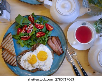 Authentic flat lay composition of a hearty morning meal. A rustic blue plate features crispy sunny-side-up fried eggs, bacon strips, grilled toast, and a fresh green salad with cherry tomatoes. Accompanied by a white teapot, fruit tea, and cutlery on a light wooden table. Perfect for cafe menus, food blogs, realistic brunch concepts, and authentic everyday lifestyle visuals.