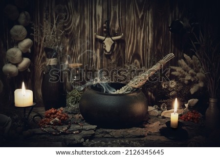 Authentic forest witch altar. Burning candles, dry herbs, iron cauldron, smoking mugwort, bulls skull mojo, bunch of garlic in wooden background in the dark, low key, selective focus.