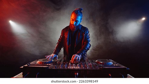 Un auténtico hipster DJ animando la fiesta. Jinete de discos con barba trabajando en una discoteca, componiendo una lista de música de baile, visto con luces rojas y azules - concepto de vida nocturna 