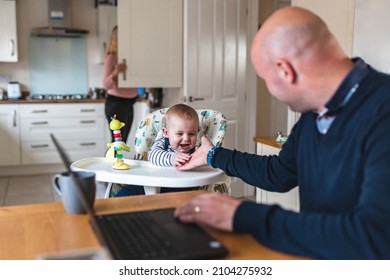 Authentic Home Shot With Young Father Working From Home And Playing With His Newborn Baby Boy In The Living Room - Concepts Of Remote Work, Children, Parenthood, Lifestyle In The UK