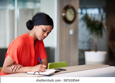 Authentic image of African American woman busy using a pen to write down her shedule that she has planned out in her notebook while sitting in the colourful business lounge.