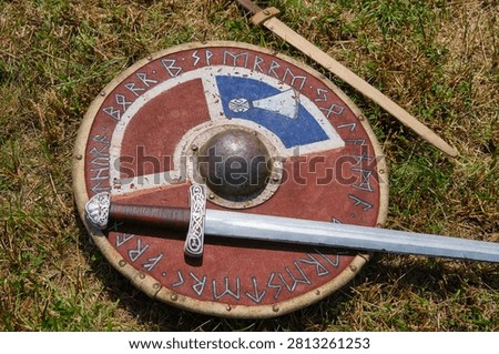 Authentic looking Viking age weaponry including a round shield and a long sword are displayed on a green grassy field for historical reenactment.