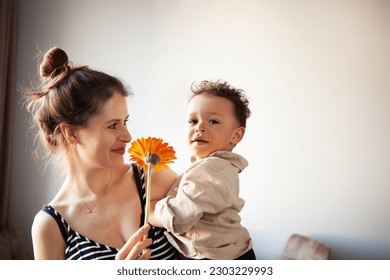 Authentic mom holding child, who gazing into camera while clutching flower. Real life and authenticity on light background. - Shutterstock ID 2303229993