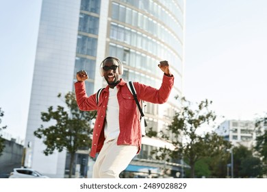 Authentic photo of happy excited young African Black man student hipster winner wearing headphones, sunglasses and backpack feeling joy having fun listening music and dancing on city street outdoors. - Powered by Shutterstock