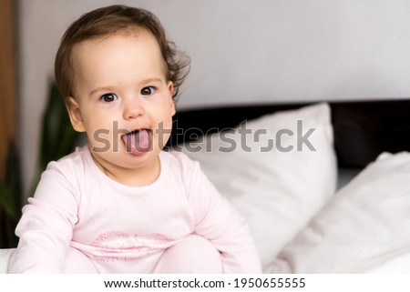 Authentic portrait cute caucasian little infant chubby baby girl or boy in pink sleepy upon waking looking at camera showing tongue in white bed. Child care, Childhood, Parenthood, lifestyle concept