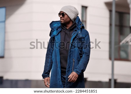 Authentic Portrait of handsome African American man walking in city, wearing stylish outfit parka coat, hoodie, knitted hat and sunglasses. street style fashion male model