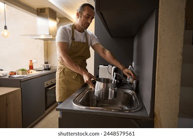 Authentic portrait of a handsome Caucasian young man in beige chef's apron, filling stainless steel saucepan with running water until it boils and cooks something in a minimalist home kitchen interior