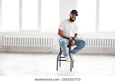 Authentic Portrait happy bearded man sitting in white loft background, smiling, wearing casual white T shirt, baseball cap and jeans, lifestyle - Shutterstock ID 2261351713