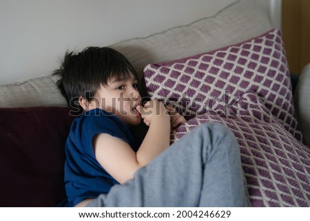 Authentic portrait Kid lying on safa biting his finger nails while watching TV, Little boy lying on couch bite his nails, Child chilling out or relaxing in living room on weekend