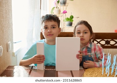 Authentic portrait of a teen boy and his younger sister holding white mockup box , sitting at table and celebrating happy life event. White gift boxes with copy space for advertising text - Shutterstock ID 2311628865