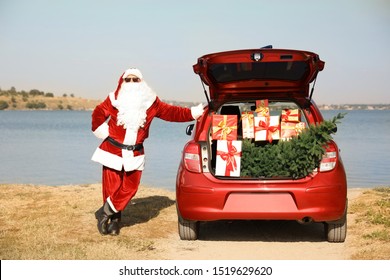 Authentic Santa Claus Near Car With Open Trunk Full Of Presents And Fir Tree Outdoors