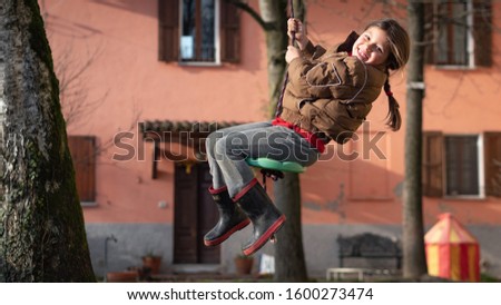 Authentic shot of cute carefree little girl is having fun on a swing outside her house in a sunny autumn day.