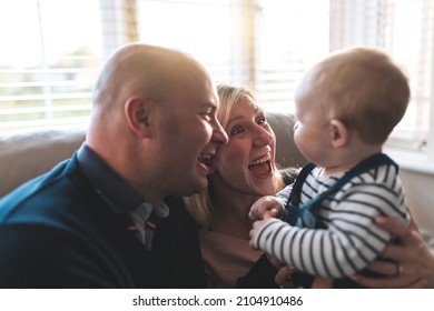 Authentic Shot Of Happy Family Portrait With Parents Laughing And Looking At Their Son While Holding Him On The Sofa At Home And Enjoying Time Together - Lifestyle And Family Concepts