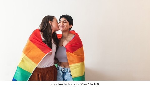 Authentic shot of happy lesbian couple wrapped in rainbow flag and giving a kiss, copy space on the right - lesbian couple at home enjoying life together