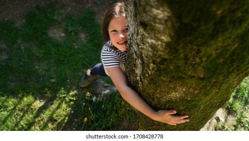 Authentic shot of a happy little girl is hugging a tree trunk and smiling in camera on a sunny day. Concept: love for nature, connection with nature, happiness, childhood, ecology, deforestation