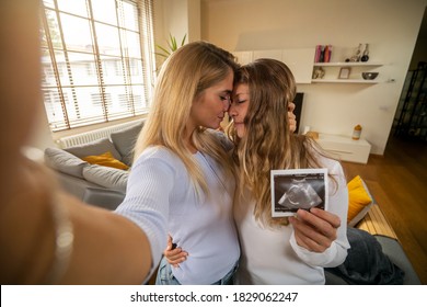 Authentic Shot Of Happy Married Homosexual Female Gay Couple Is Making Video Technology Call To Friends Or Family With Smartphone From Home And Showing Ultrasound To Announce Their Future Child Birth.