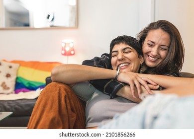 Authentic shot of happy married homosexual female gay couple laughing and embracing on the sofa with rainbow pride flag on background - lesbian couple at home enjoying life together - Shutterstock ID 2101856266
